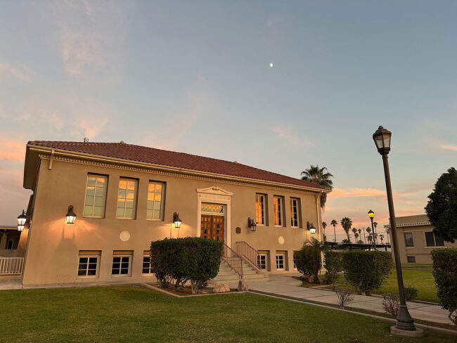 A stucco library at sunset. Hedges and a lampost are visible.