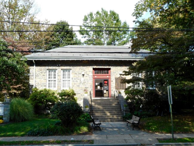 A rectangular stone library with red trimmed door and trees and shrubs around it.