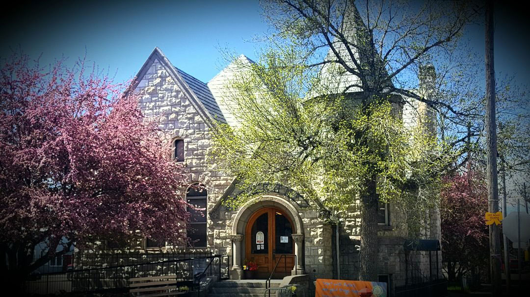 A stone building with wooden doors, blocked by a pink blooming tree and green tree. The building has pointed roofs and a turret.