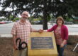A man and a woman stand holding hands before a plaque commemorating the Eastside Library. One holds a pair of giant scissors.