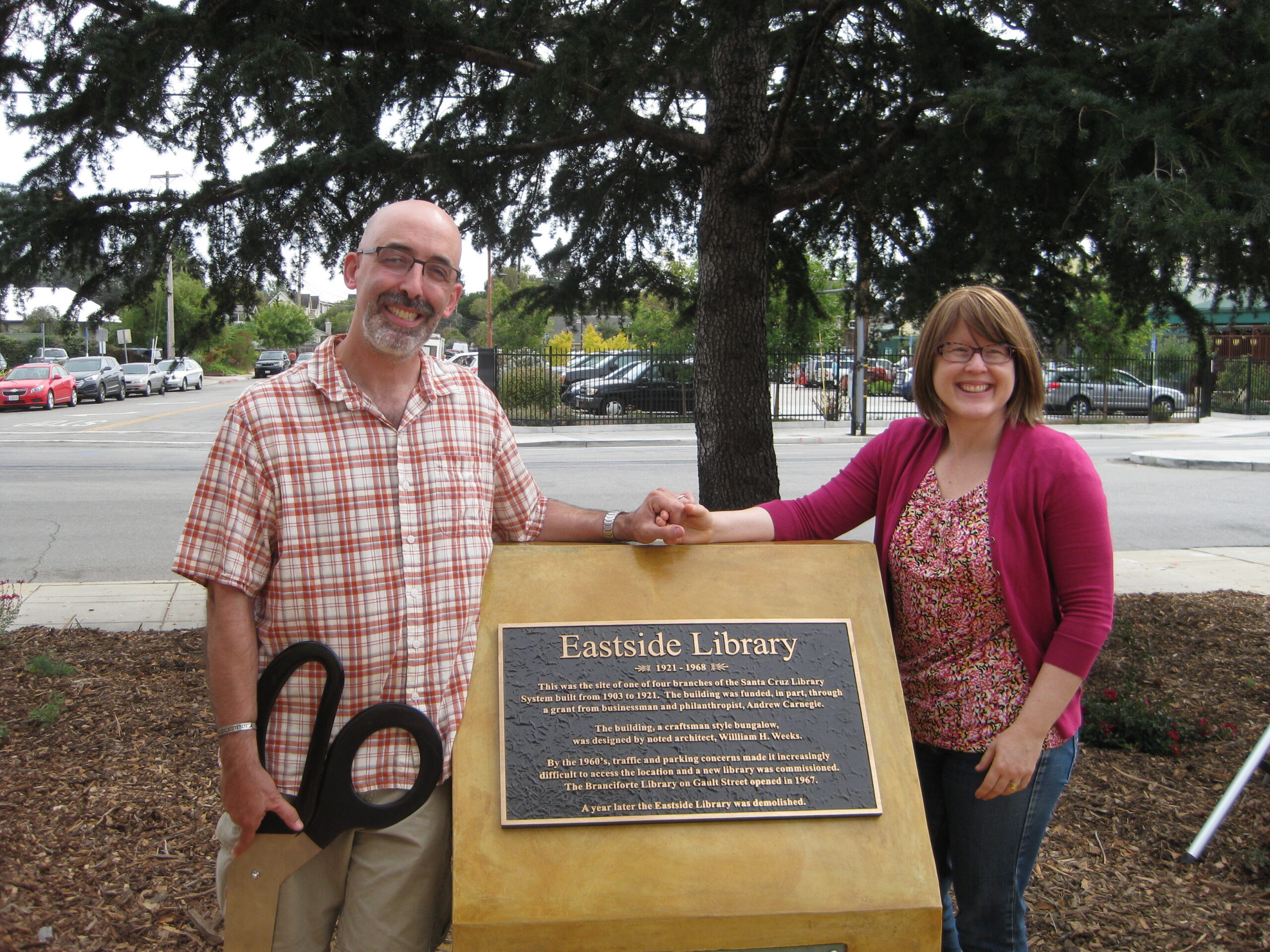 A man and a woman stand holding hands before a plaque commemorating the Eastside Library. One holds a pair of giant scissors.