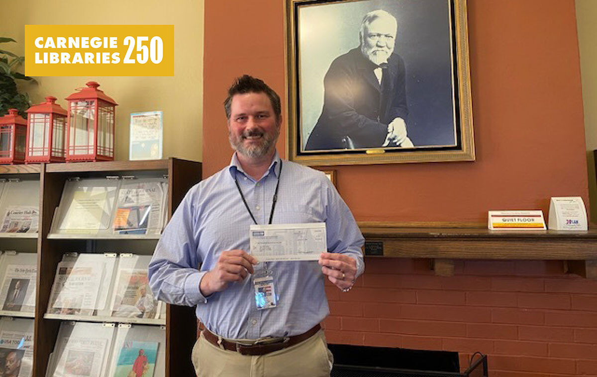 A man stands before a photo of Andrew Carnegie holding a check.